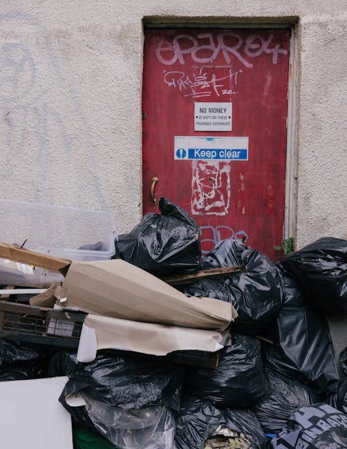 A pile of black garbage bags and assorted discarded items, including a flattened cardboard box and a piece of white foam, are stacked against the base of a red metal door set into an exterior wall with a rough, light-colored surface. The door is covered in graffiti tags and features two posted signs: one reading 'No Money - Keep Clear' in black and white, and another blue sign with white text that says 'Keep Clear.' The surrounding environment appears to be an alley or service area with minimal visible context, emphasizing the rubbish accumulation intended for collection or disposal outside a building. The lighting is natural, casting soft shadows and highlighting the textures of the bags, the graffiti, and the door's weathered finish. This scene exemplifies typical private waste handling or on-site clearance scenarios that Waste Disposal Islington may service, illustrating the clutter that can accumulate in urban settings requiring rubbish removal services.