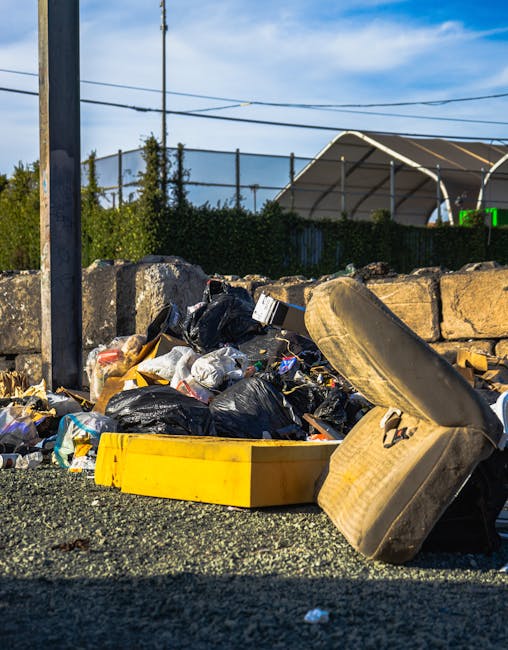 An outdoor scene showing a large pile of mixed waste and rubbish bags accumulated on the ground near a stone wall and a metal pole. The waste includes black plastic bin bags, cardboard boxes, and various debris scattered on a gravel surface. A worn, used car tire with visible tread and a beige, textured interior lining is leaning against the rubbish pile. In the background, there are green shrubbery, a tall chain-link fence, and a sports field with a shelter structure made of metal and fabric. Overhead, there are power lines and a partly cloudy sky with blue patches, indicating daytime. The scene suggests an unmanaged or improperly disposed of waste area, highlighting the importance of commercial rubbish removal services to keep outdoor environments clean, like those provided by Waste Disposal Islington, for proper waste handling and alternative disposal options beyond regular collection.