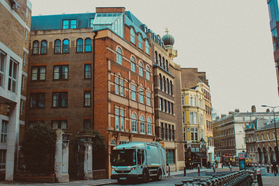 A commercial street scene showing multiple multi-storey buildings with varied architectural styles, including red-brick and yellow-painted facades, set closely together along a paved urban road. In the foreground, a white garbage collection truck is parked adjacent to a sidewalk, appearing ready to perform waste collection or rubbish removal. The truck's surface is smooth with a practical design, and it has a large cab with tinted windows. To the left and right of the truck, there are metal bike racks and lampposts lining the street, signifying an urban environment. The buildings feature numerous windows, some with arched top sections, and decorative architectural elements such as cornices and window frames, with a domed tower visible on one of the structures. The lighting suggests a daytime setting with evenly diffused natural light, and there are no visible pedestrians or other vehicles, emphasizing an area suited for private or commercial rubbish collection services. The scene reflects a typical city block where waste disposal or clearance activities, potentially handled by Waste Disposal Islington, support ongoing urban maintenance and rubbish removal efforts.