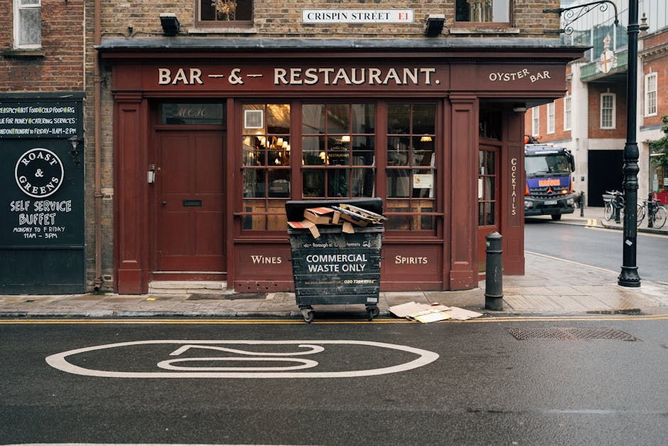 A black wheeled waste container labeled 'Commercial Waste Only' is positioned on the pavement directly in front of a red-painted building housing a bar and restaurant, with large glass windows displaying warm interior lighting. The container is overflowing with flattened cardboard boxes, some spilling onto the ground, and appears to be used for ongoing waste collection related to the establishment. The surrounding environment includes a lamppost on the right, a black bollard adjacent to the container, and a parked truck further down the street behind the building. The street surface features a white marking indicating a bus stop, and the scene is illuminated by natural daylight, suggesting daytime hours. The setting exemplifies a typical urban commercial area where private waste disposal services, such as those provided by Waste Disposal Islington, are employed for on-site rubbish collection outside hospitality premises, with visible waste management practices supporting business needs.