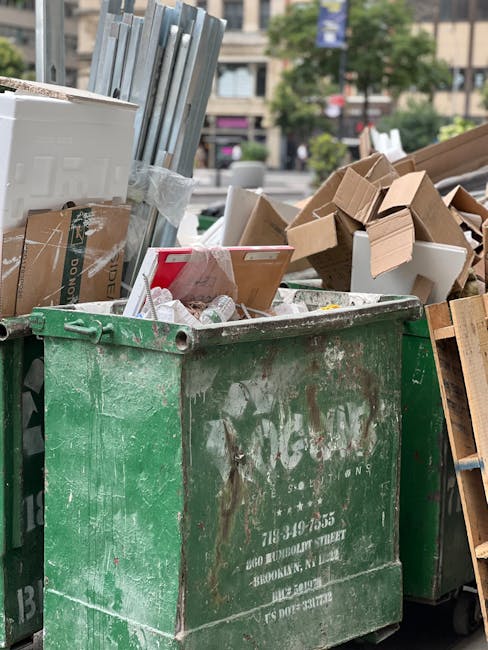 A large, weathered green skip positioned on a paved surface in an outdoor urban environment, filled with mixed waste including flattened cardboard boxes, some partially open or crushed, and a few white plastic bottles scattered inside. Adjacent to the skip are additional discarded materials such as flattened cardboard sheets, small wooden pallets, and a stack of unassembled metal or plastic fencing panels leaning against the side of the skip. The background shows a row of trees and multi-storey buildings, with cars and pedestrians visible in the distance, indicative of a city setting. This scene illustrates a typical on-site waste collection for bulk rubbish removal, where local residents or businesses may do private disposal, aligning with alternative rubbish handling methods outside council curbside services. The overall atmosphere suggests an active waste management effort, with the disposal unit clearly marked and used for clearing large amounts of refuse in a public or commercial area. Waste Disposal Islington is likely responsible for the collection, supporting effective clearance of bulk residential or commercial waste.