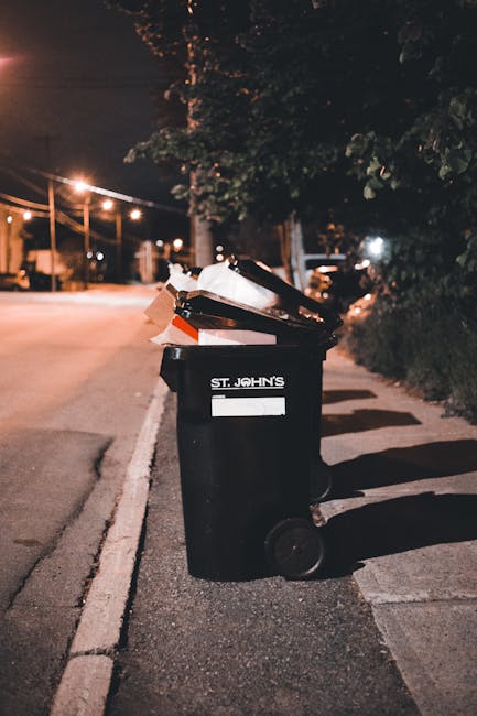 A black wheeled rubbish bin labeled 'St. John’s' is positioned on a sidewalk near the edge of the road during nighttime. The bin is overflowing with various waste materials, including cardboard boxes, plastic bags, and paper, some of which are leaning or protruding from the open lid. The surrounding environment includes dark trees and shrubbery along the pavement, with a row of streetlights casting warm, diffuse illumination across the scene. In the background, blurred vehicles and distant light sources are visible, suggesting an urban or suburban residential area. The scene emphasizes the importance of designated waste disposal and independent rubbish collection in non-urban settings, aligning with services provided by Waste Disposal Islington for private waste handling. The lighting and setting reflect typical conditions for evening rubbish disposal, illustrating the need for reliable rubbish removal services to manage overflowing bins and maintain cleanliness outside residential properties.