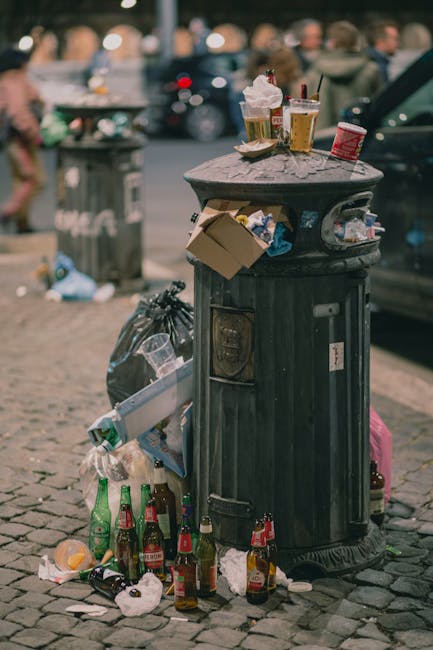A tall, rectangular blue rubbish bin with a closed lid, positioned on a grassy area outdoors. The bin is made of durable plastic with a smooth finish and has a small handle on top for opening. A yellow sticker with the phrase 'FIGHT BACK!' is applied across the bin's front, slightly torn at the edges. To the left of the blue bin, part of a red container with black dots on its surface is visible, and it appears to be made of metal or plastic. The red container has a slightly domed, metallic lid and is placed on the pavement or ground nearby. Behind the bins, a metal pole supports a small blue sign, partially visible at the top left. The surrounding environment includes a bike lane or pathway with metal barriers or posts along a roadside, and the background shows a bright sky with soft sunlight, creating a natural scene related to waste disposal and rubbish collection in an outdoor urban area.