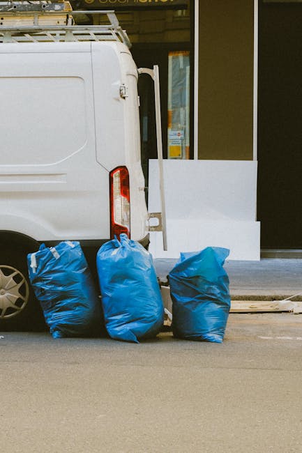 Four large blue rubbish bags filled with waste are positioned on the pavement beside a white commercial van with a modern design, featuring a vertical tail light on the left side and a visible rear door. The bags are made of thick plastic, slightly crumpled, and appear to be filled with household or construction debris. The scene is set in an urban environment, with the van parked close to the entrance of a building that has a glass window and door, partially visible behind the bags. The pavement surface is smooth and clean, with a curb separating it from the asphalt road. Behind the bags, there is a small section of a white structure or barrier, possibly an outdoor step or low wall, and some reflections on the building's glass suggest nearby structures or signage. This setup implies an activity related to independent waste disposal or on-site rubbish collection, similar to what might be arranged through private waste management services such as Waste Disposal Islington, to handle accumulated waste outside commercial or residential premises.