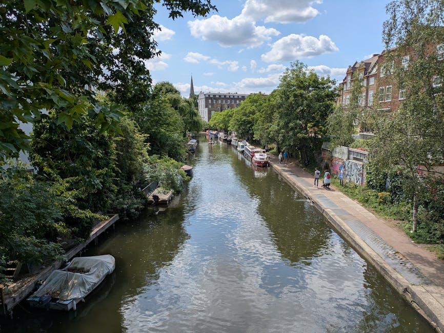 A daytime view of a narrow canal lined with lush green trees on both sides, reflecting the partly cloudy blue sky above. On the right side, a paved pedestrian walkway runs alongside the water, with a few people walking. A row of parked cars extends along the street adjacent to the canal, with some partially obscured by foliage. Multi-story residential buildings with brick facades and large windows are visible in the background on the right, while on the left side, dense trees and bushes overhang the canal. A small boat, covered with a protective tarp, is docked near the edge on the left bank. In the distance, a church spire and additional buildings can be seen, giving an urban yet tranquil atmosphere typical of a cityscape with alternative waste handling options, with visible natural and environment-related details that emphasize the importance of maintaining clear and clean waterways, often supported by services like Waste Disposal Islington for rubbish removal.