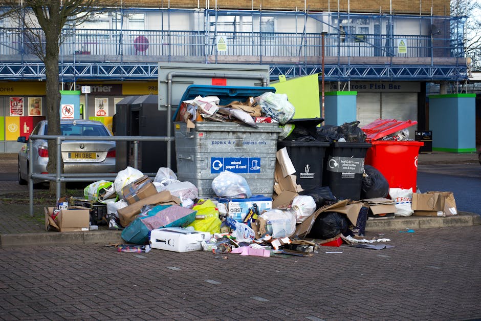 A large pile of assorted outdoor waste is accumulated on a paved area next to a metal railing, consisting of overflowing cardboard boxes, plastic bags full of rubbish, crumpled paper, and discarded packaging. The contents include brown and white cardboard boxes, some flattened and others still intact, along with black and green plastic trash bags, all spilling onto the pavement. A large grey wheelie bin with a blue recycling sticker labeled for mixed paper and card is positioned centrally, with its lid open and overflowing with paper waste. To the right, a black recycling bin and a bright red disposal bin are placed side by side, also filled with refuse. Behind the bins, there is a multi-story building with a blue metal scaffolding structure and several closed shopfronts with signs, indicating a commercial area. The scene is lit by natural daylight, with an overall impression of an uncollected rubbish accumulation that local waste management services might handle through private or independent collection options, such as those offered by Waste Disposal Islington in alternative waste removal contexts.