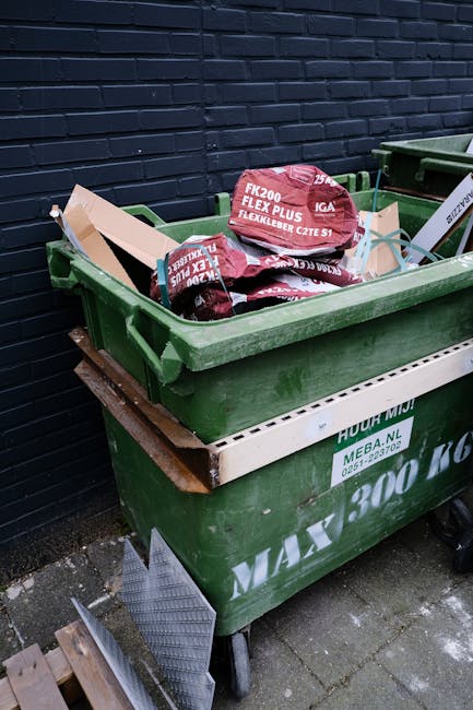 A green wheelie bin filled with red and black flexible hoses, some branded with text indicating product specifications, is positioned on a paved area outdoors against a dark blue brick wall. The bin is partially supported by a white platform or liner inscribed with waste disposal information and the logo of Waste Disposal Islington. The hoses inside appear to be coiled or bent, with some overlapping and protruding above the rim of the bin. To the side, a small section of another similar green bin is visible. The surrounding environment suggests a back alley or service area, typical for private waste collection or bin storage for rubbish removal services. The atmosphere is illuminated by natural light, highlighting the textures of the plastic hoses and the rough surface of the brick wall, emphasizing the scene's functional aspect of refuse management and recycling support.