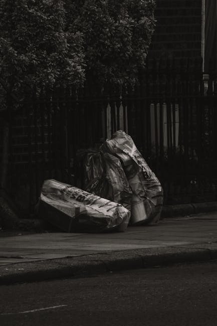 A large pile of assorted outdoor waste is accumulated on a paved area next to a metal railing, consisting of overflowing cardboard boxes, plastic bags full of rubbish, crumpled paper, and discarded packaging. The contents include brown and white cardboard boxes, some flattened and others still intact, along with black and green plastic trash bags, all spilling onto the pavement. A large grey wheelie bin with a blue recycling sticker labeled for mixed paper and card is positioned centrally, with its lid open and overflowing with paper waste. To the right, a black recycling bin and a bright red disposal bin are placed side by side, also filled with refuse. Behind the bins, there is a multi-story building with a blue metal scaffolding structure and several closed shopfronts with signs, indicating a commercial area. The scene is lit by natural daylight, with an overall impression of an uncollected rubbish accumulation that local waste management services might handle through private or independent collection options, such as those offered by Waste Disposal Islington in alternative waste removal contexts.