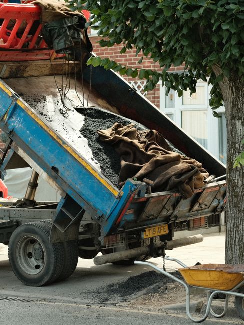 A small blue and yellow articulated tipper truck with a black asphalt load in its open rear container, positioned on a paved street near a tree and a brick building. The truck's rear is tilted upward as the asphalt is being offloaded, with a brown cloth or tarp partially covering the material. To the right of the truck, a yellow wheelbarrow rests on the pavement, and a red plastic crate is placed on top of the truck's side. The scene appears to be part of outdoor construction or road maintenance work, indicative of private waste or material disposal services, with greenery and a residential building visible in the background. The environment is well-lit, suggesting daylight, and the presence of tools and equipment underscores ongoing site clearing activities, relevant to rubbish removal and disposal processes overseen by Waste Disposal Islington.