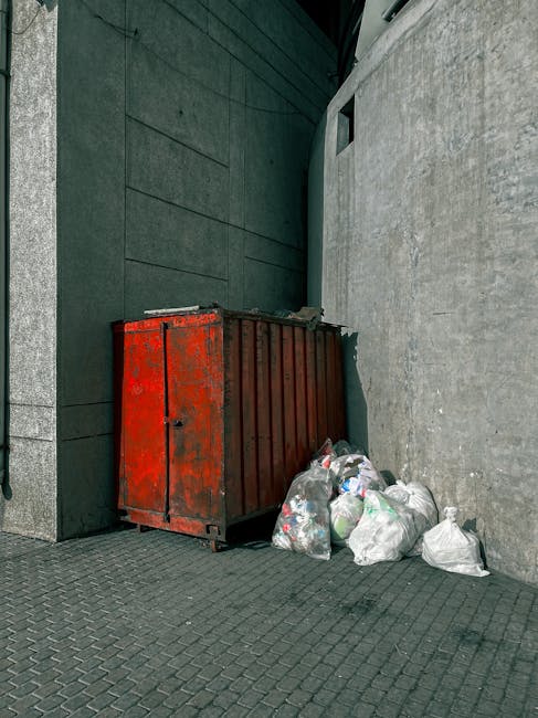 A large, red metal waste skip with visible rust and wear sits against a dark green wall and a light grey concrete wall in an urban outdoor setting. The skip is positioned on a paved surface composed of small, dark grey interlocking bricks. Several clear and white plastic bags filled with assorted rubbish are piled next to the skip, some leaning against its side. The background features a modern building façade with smooth, dark green panels and a vertical black strip, along with a small rectangular window or vent near the top of the green wall. The environment appears clean but cluttered with waste, suggesting a private collection or on-site disposal scenario typical of waste management services like Waste Disposal Islington, which handle rubbish removal and alternative waste handling solutions in urban areas. The natural lighting is diffuse, with no strong shadows, emphasizing procedural waste collection at a commercial or residential site.