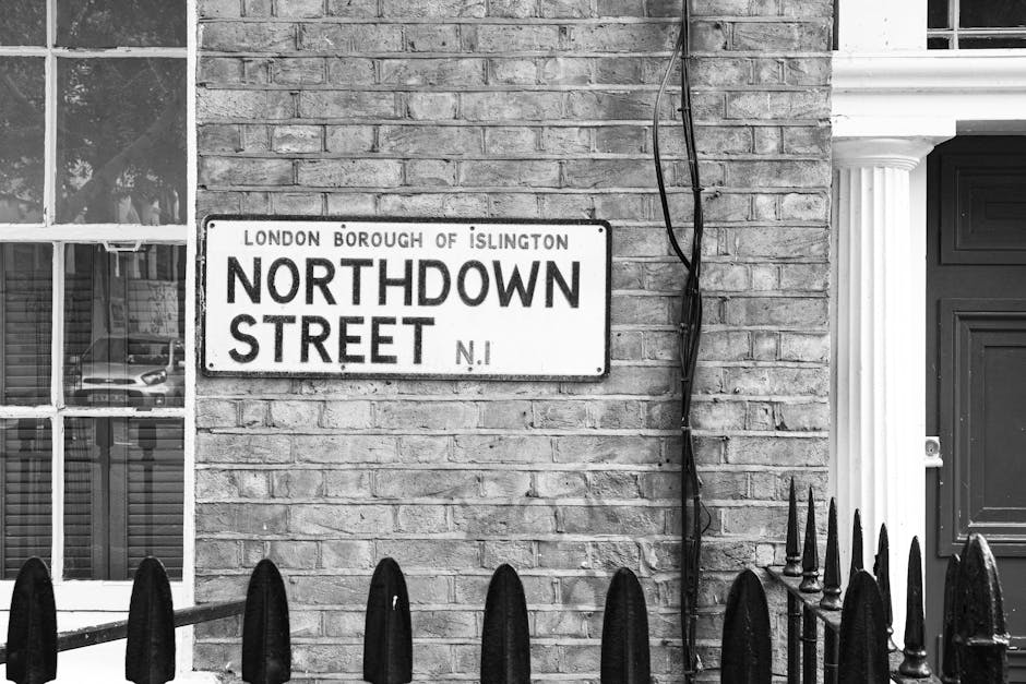 A black-and-white photograph depicts a residential street scene featuring a brick wall with a London Borough of Islington street sign reading 'Northdown Street N1' attached to it. To the left, a portion of a window with multiple panes reflects parked cars and nearby buildings. To the right of the sign, exposed electrical wires run vertically down the brick wall, secured by black clips. The lower part of the image shows a decorative black metal fence with pointed finials in the foreground, separating the street from the property. Adjacent to the brick wall, a section of a wooden front door painted dark green or black is partially visible, framed by white classical architectural columns. The environment appears well-maintained and typical of a London residential area, providing an appropriate context for discussing alternative waste handling or on-site clearance services such as those offered by Waste Disposal Islington in relation to local rubbish removal needs.